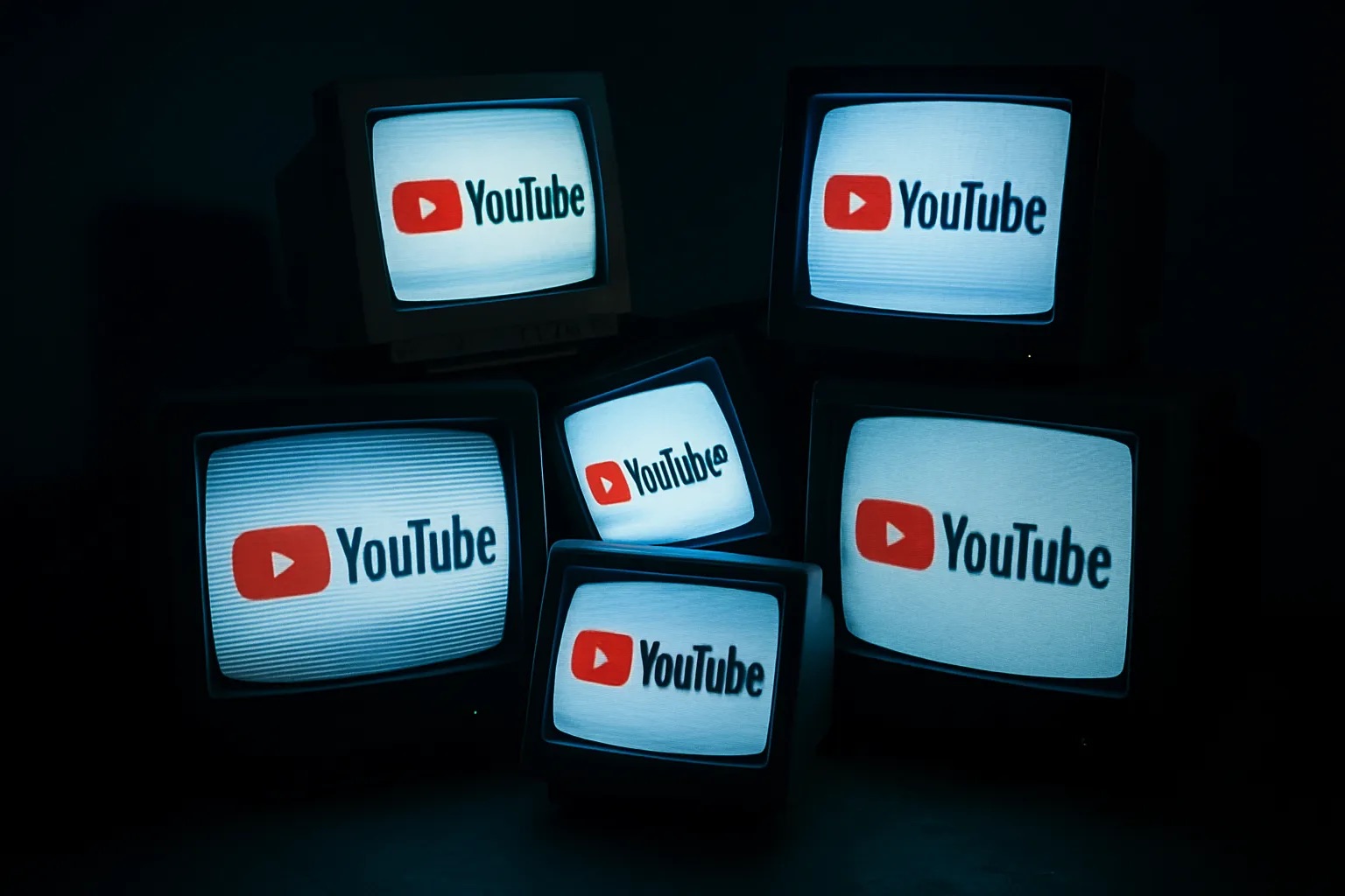 Varios monitores CRT antiguos apilados de forma desordenada en una habitación oscura, cada uno mostrando el logo de YouTube con líneas de barrido visibles, evocando una atmósfera retro y nostálgica que representa el concepto de YouTube Premium Lite sin anuncios.
