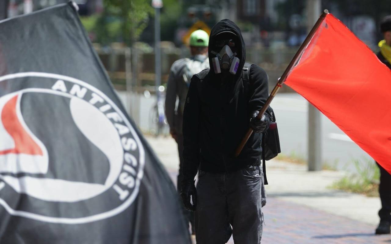 Manifestante encapuchado con máscara antigás sostiene una bandera roja junto a otra con el símbolo de Antifa, en el contexto de protestas en Estados Unidos tras la designación de Antifa organización terrorista