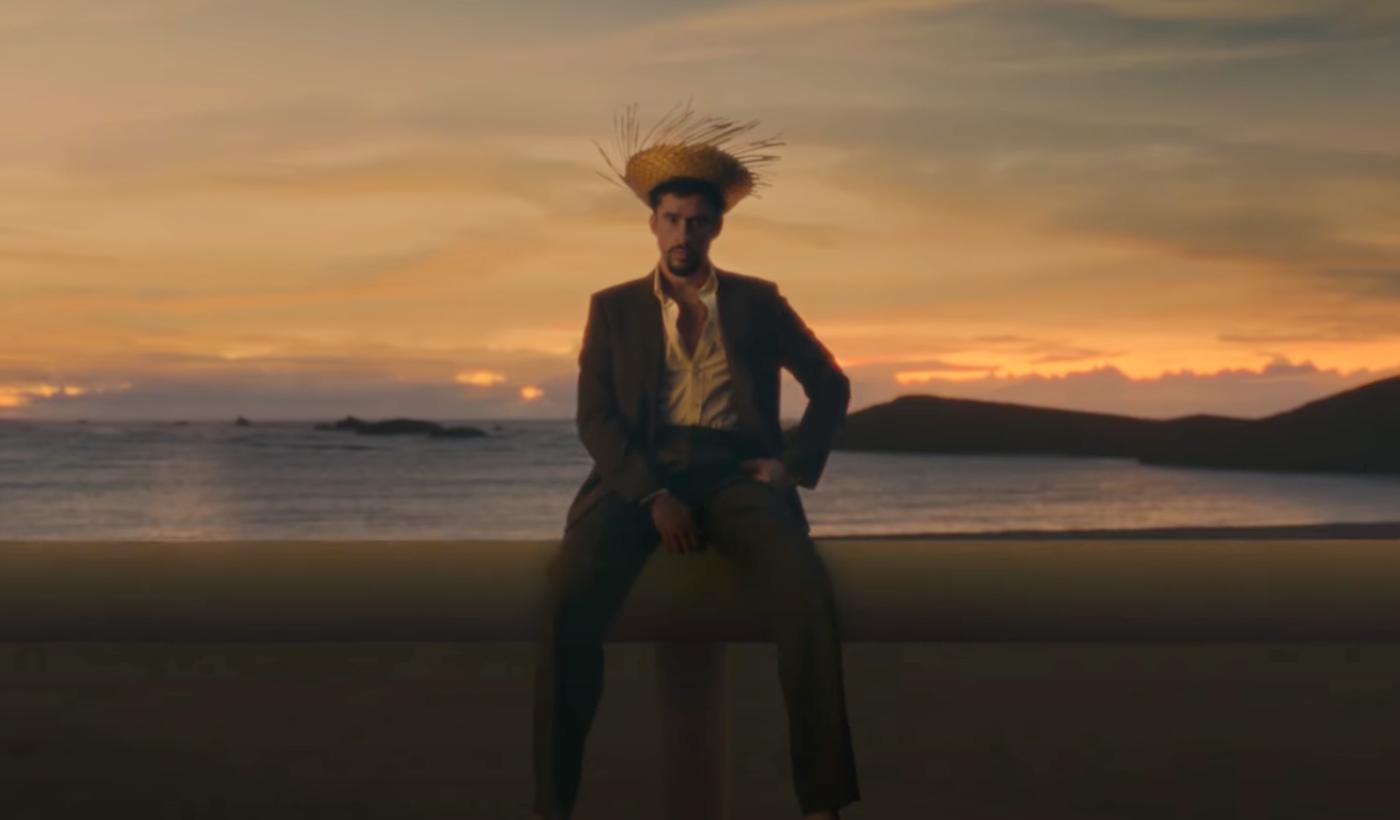 Bad Bunny sentado sobre un arco de fútbol americano con traje oscuro y sombrero de paja, frente a un atardecer junto al mar