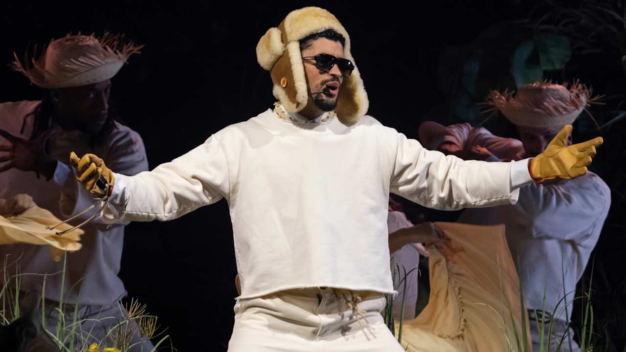 Bad Bunny vestido de blanco con gorro de invierno y guantes amarillos, cantando en el escenario junto a bailarines con sombreros de paja