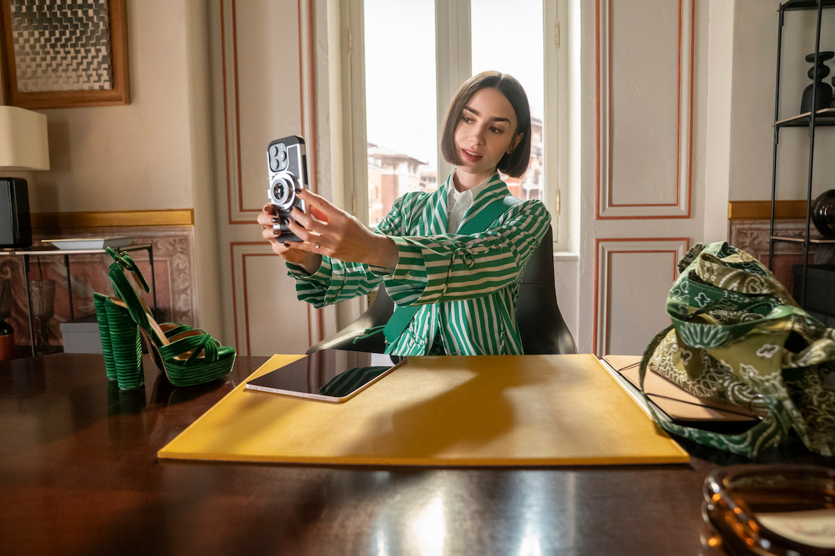 Lily Collins como Emily Cooper tomando una selfie en su oficina en Roma durante Emily en París temporada 5, simbolizando su nueva etapa profesional entre París y Roma