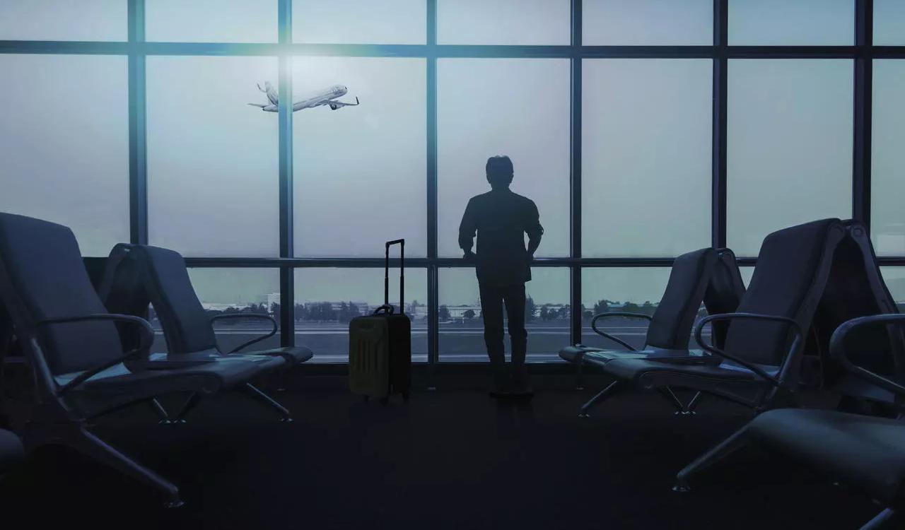 Persona observando por una ventana de aeropuerto mientras un avión despega, con su maleta junto a los asientos vacíos, en un contexto asociado a la cancelación de vuelos por actualización