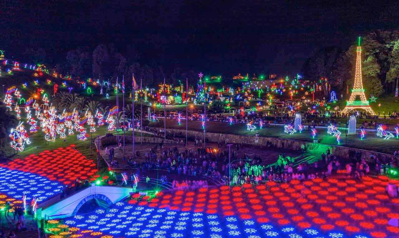 Vista panorámica nocturna del alumbrado del Puente de Boyacá, con figuras navideñas iluminadas, estructuras de colores en el suelo y una torre luminosa similar a la Torre Eiffel, en medio de visitantes recorriendo el lugar durante el alumbrado del Puente de Boyacá