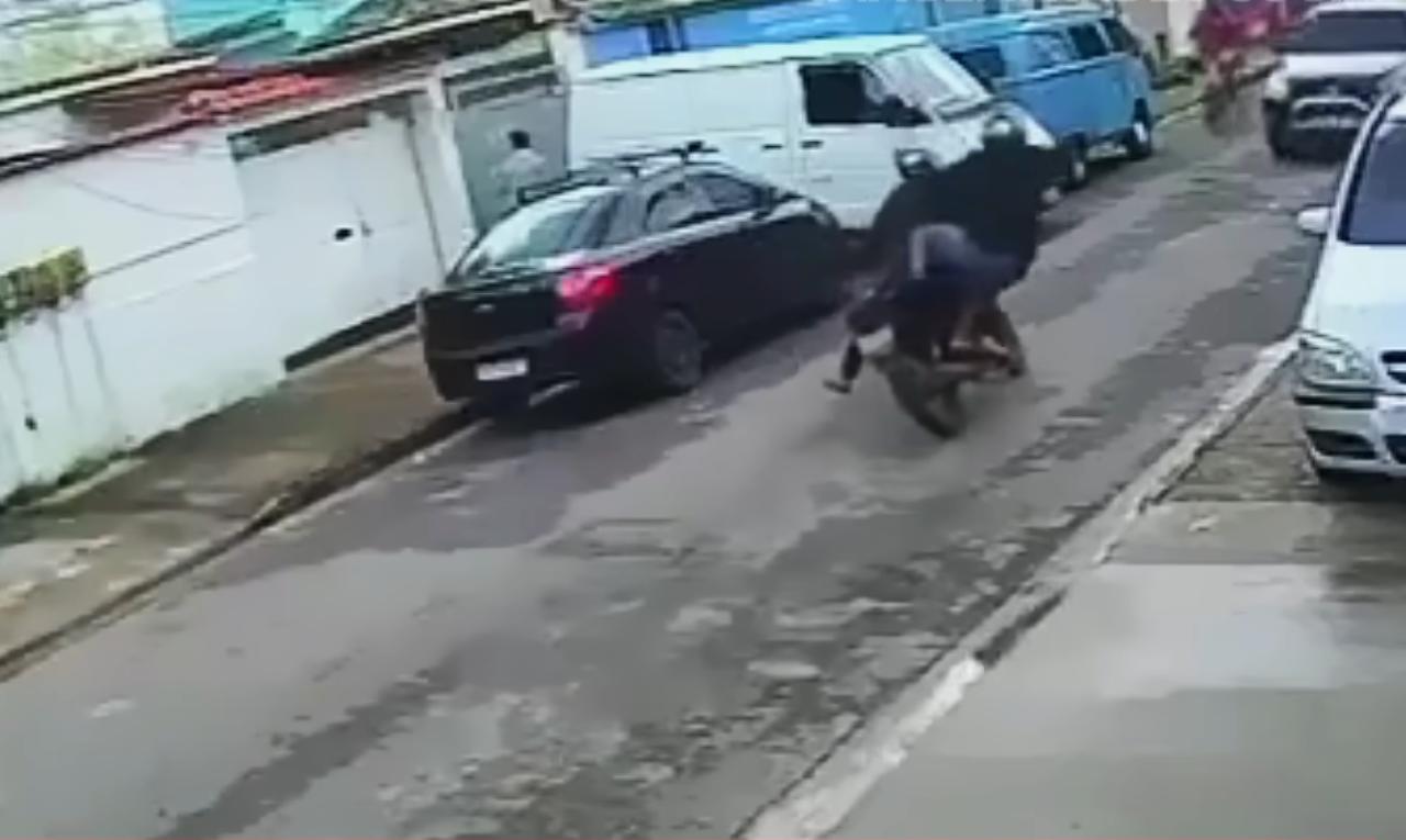La imagen muestra a dos hombres en una motocicleta, donde el acompañante está saltando del vehículo en movimiento, extendiendo una pierna hacia adelante para bajarse justo frente a una camioneta blanca. La moto sigue avanzando mientras el hombre intenta tocar el suelo, aparentemente para acercarse al vehículo o interceptarlo. La escena ocurre en una calle residencial estrecha, con vehículos estacionados a ambos lados y captada por una cámara de seguridad.