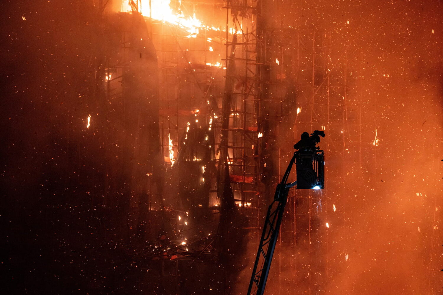 Un bombero en una plataforma elevadora intenta controlar el incendio en Hong Kong frente a una fachada envuelta en llamas y andamios de bambú, mientras chispas y fragmentos incandescentes llenan el aire durante la noche