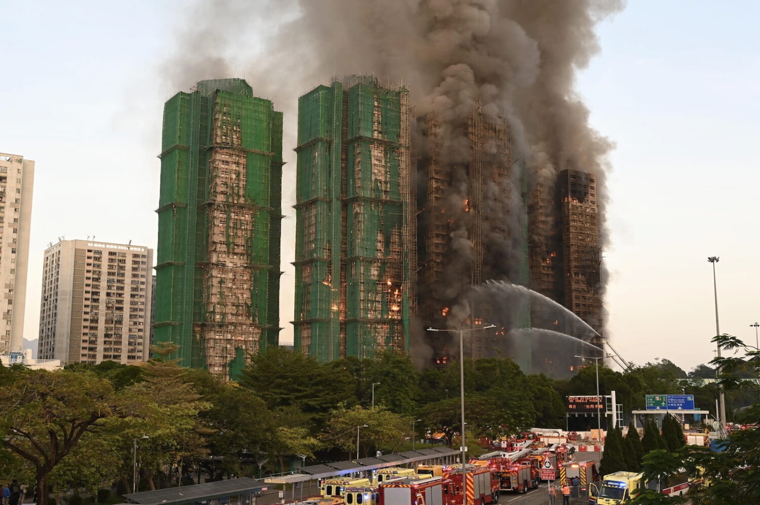 Vista diurna del incendio en Hong Kong, con varias torres envueltas en andamios de bambú y cubiertas por densas columnas de humo, mientras camiones de bomberos y equipos de emergencia trabajan desde el suelo para contener las llamas