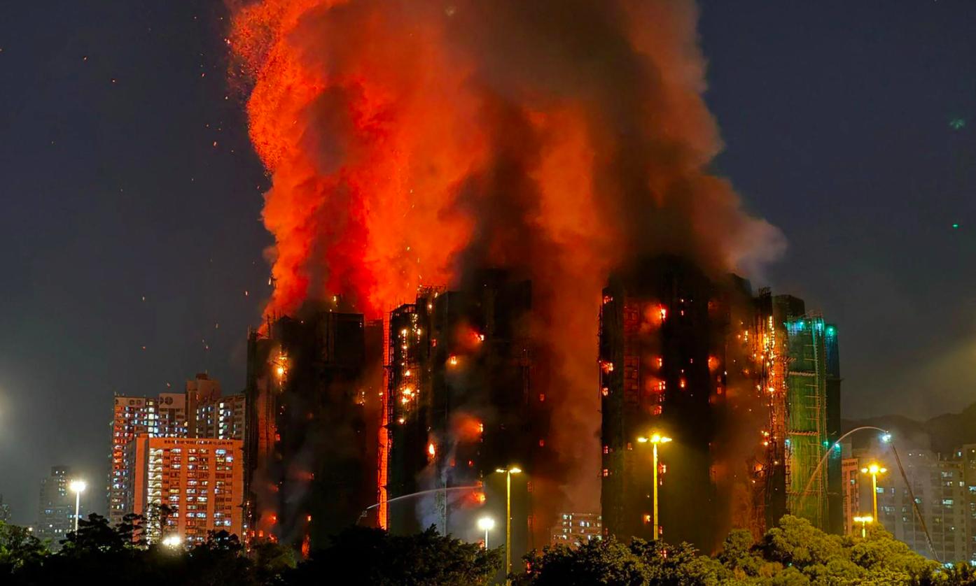Vista nocturna del incendio en Hong Kong, con varias torres residenciales envueltas en llamas intensas y una columna de humo naranja que asciende sobre los andamios de bambú, mientras luces urbanas iluminan la escena desde el suelo