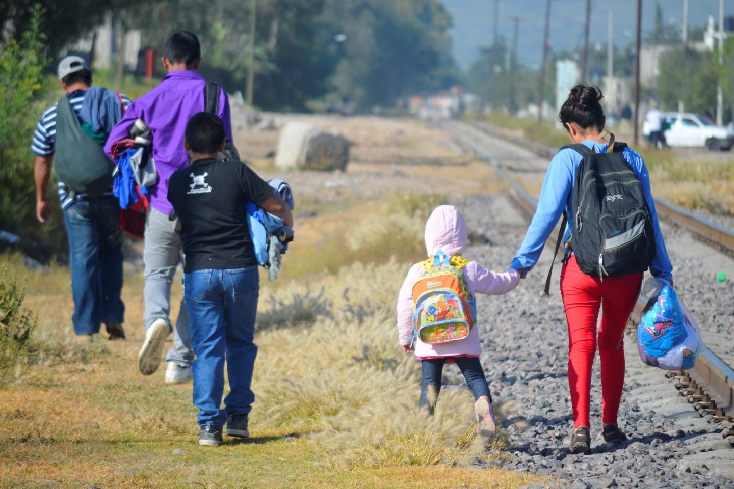 Familias caminando junto a una vía férrea con mochilas y bolsas, simbolizando la migración de países del Tercer Mundo a Estados Unidos, escena vista desde atrás en un trayecto rural