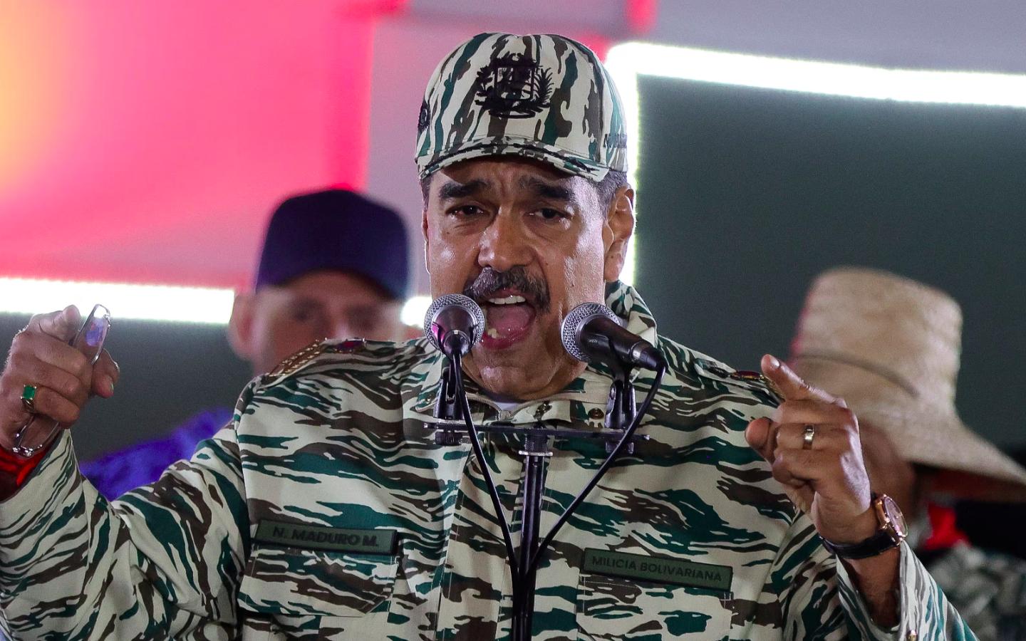Nicolás Maduro aparece con uniforme militar y gorra de camuflaje mientras ofrece un discurso en un escenario iluminado, sujetando micrófonos con ambas manos, en una imagen relacionada con la designación terrorista del Cartel de los Soles