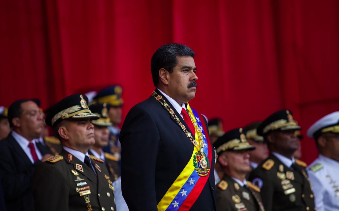 Nicolás Maduro aparece de pie durante un acto oficial rodeado de altos mandos militares venezolanos, con una banda presidencial y un fondo rojo intenso, en una imagen relacionada con la designación terrorista del Cartel de los Soles