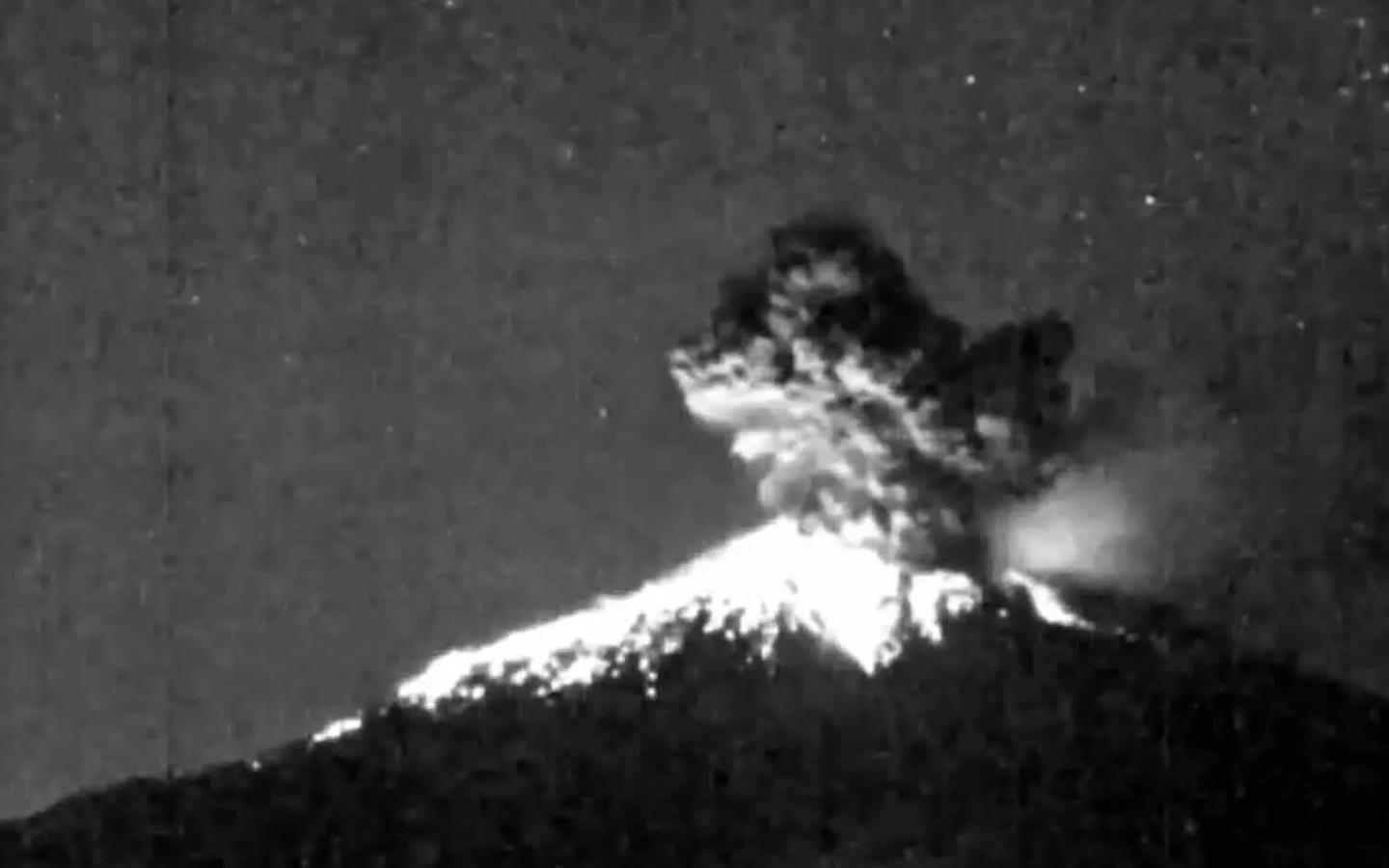 La imagen muestra una erupción nocturna del volcán Sakurajima captada por una cámara de vigilancia en blanco y negro. Se ve un chorro de material incandescente ascendiendo desde el cráter, iluminando las laderas superiores del volcán. Sobre la cúspide aparece una nube densa de ceniza y gases que se eleva con una forma irregular, empujada por la fuerza de la explosión. Alrededor, el cielo oscuro sirve de contraste y resalta el brillo volcánico, mientras pequeños puntos luminosos —posiblemente estrellas o ruido de la cámara— completan la escena. La sensación general es de fuerza súbita, un destello violento en plena noche que revela la intensidad de la actividad eruptiva.
