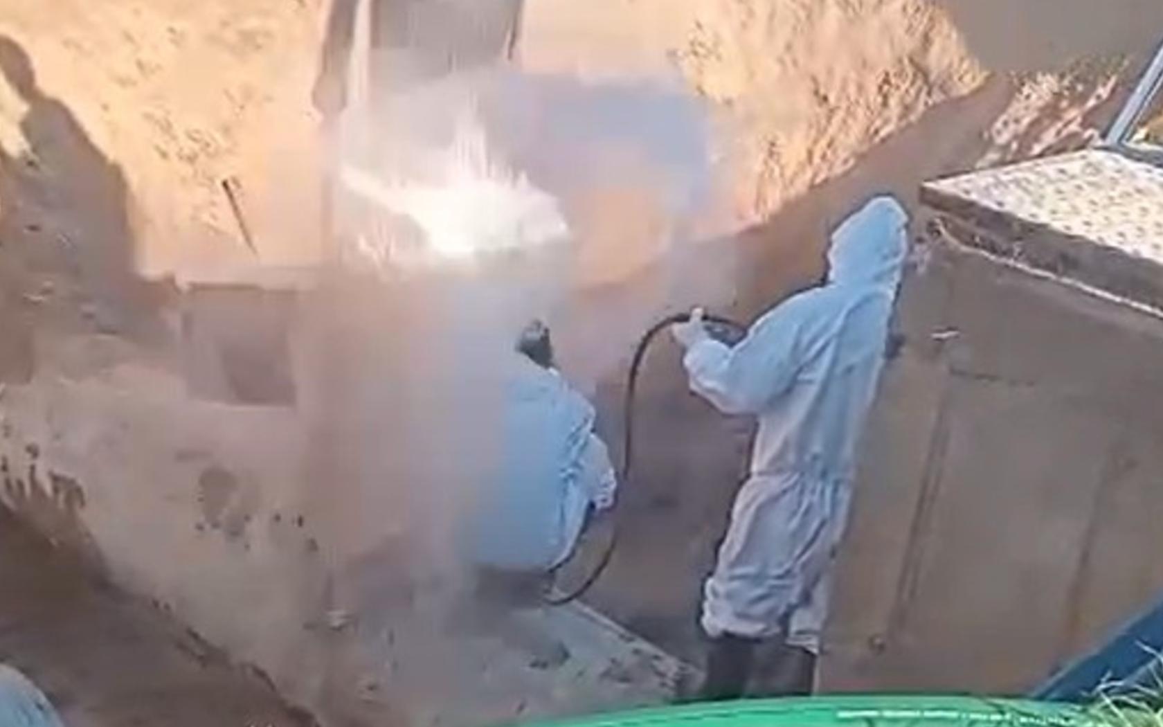 Dos trabajadores con trajes protectores blancos se encuentran dentro de una zanja mientras realizan tareas sobre una tubería. En el centro de la escena, un chorro de agua a presión emerge repentinamente, generando una nube blanca que los envuelve. Uno de los operarios está agachado y el otro sostiene una manguera, ambos sorprendidos por la explosión de agua. El entorno muestra paredes de tierra y equipamiento de obra