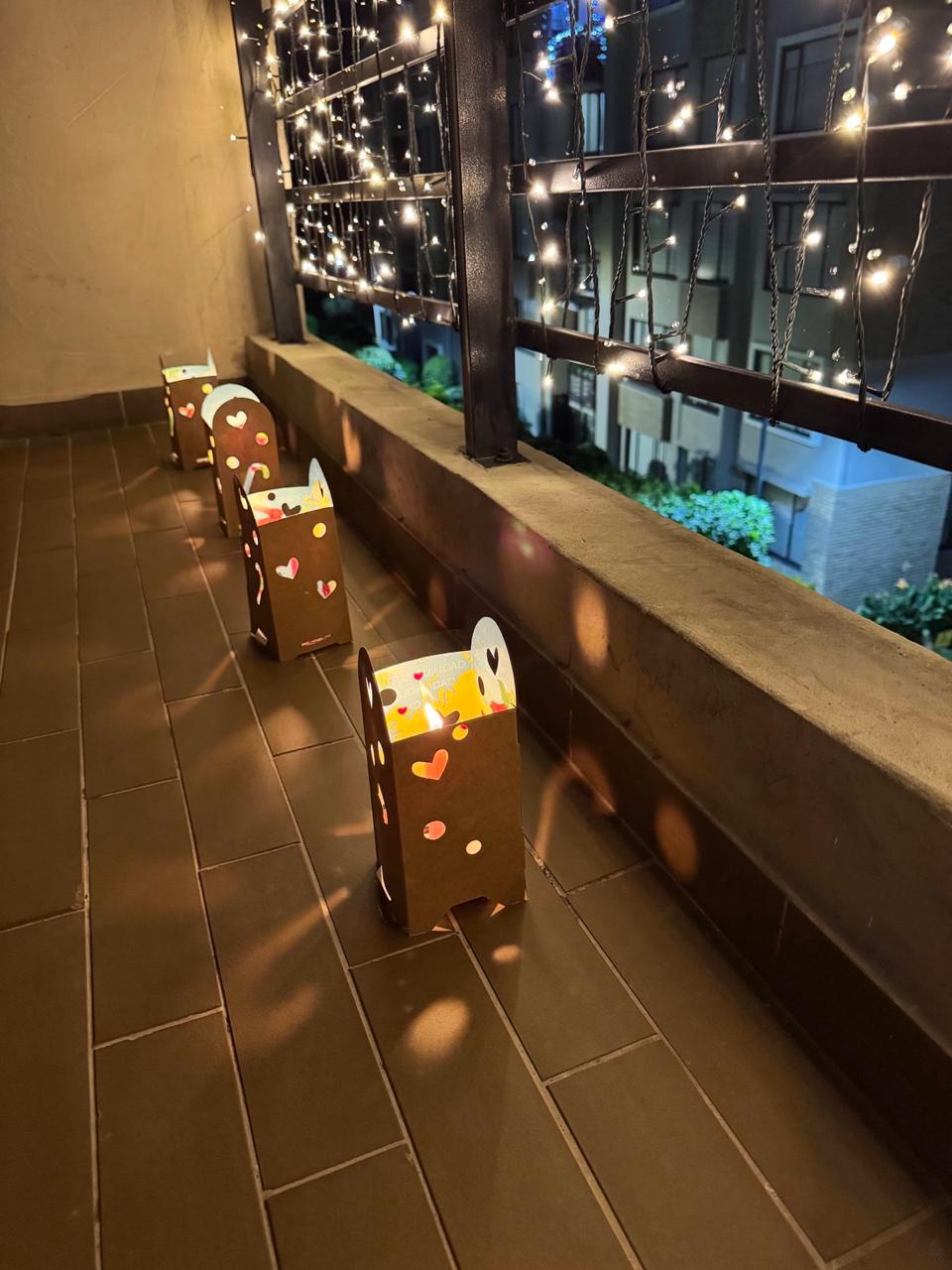 Faroles de papel iluminados sobre el piso de un balcón decorado con luces navideñas durante el Día de las Velitas, proyectando reflejos cálidos en las baldosas mientras se observa un conjunto residencial al fondo