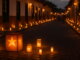 Una calle colonial colombiana iluminada al anochecer con velas y faroles durante el Día de las Velitas, mostrando una fila de luces cálidas a lo largo de las fachadas y un ambiente festivo y tradicional