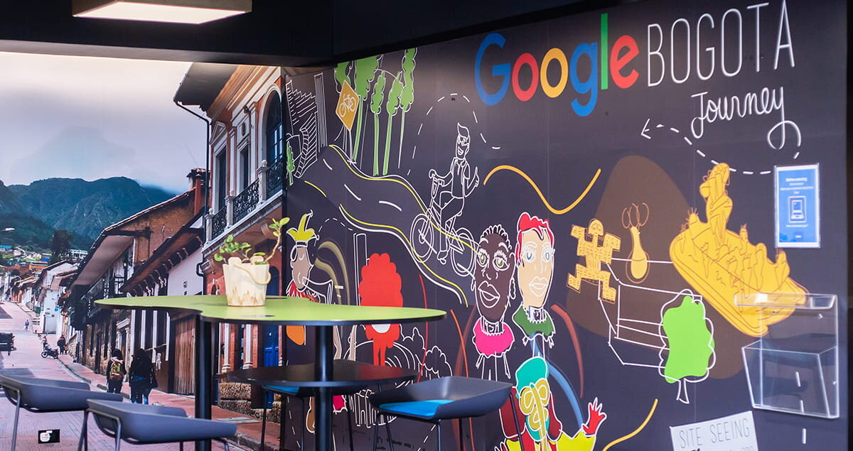Interior de la oficina de Google en Bogotá, con una pared decorada con ilustraciones coloridas y la palabra ‘Google Bogotá’, junto a una fotografía de una calle tradicional con montañas al fondo