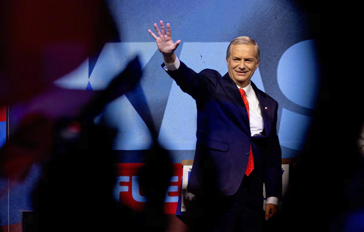 José Antonio Kast saluda con la mano levantada durante un acto político en Chile, vestido con traje oscuro y corbata roja, ante un escenario con su nombre de fondo y público al frente