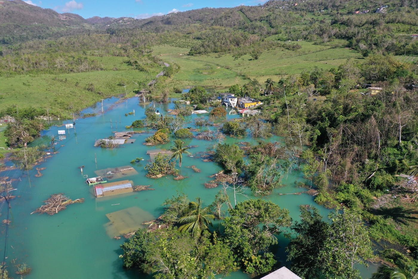 Vista aérea de una zona rural del Caribe severamente inundada tras un huracán; casas sumergidas, árboles caídos y caminos cubiertos por agua turquesa, mostrando el tipo de desastre que programas de UNESCO buscan documentar y comunicar con mayor precisión