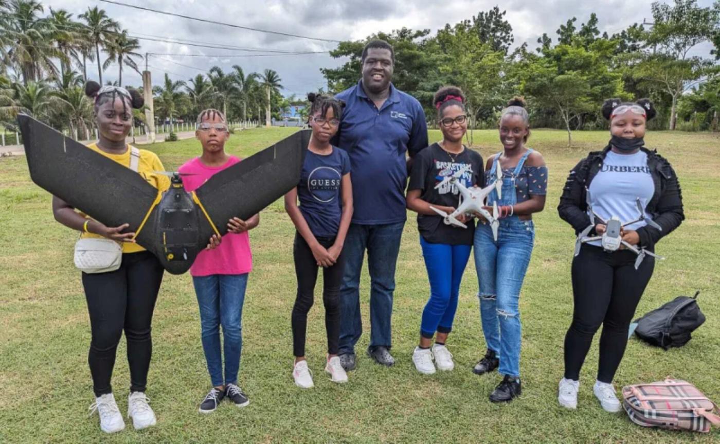 Grupo de jóvenes participantes en una capacitación apoyada por UNESCO posando al aire libre junto a diferentes tipos de drones, incluyendo un dron de ala fija y modelos multirrotor, mostrando su entrenamiento tecnológico en el Caribe